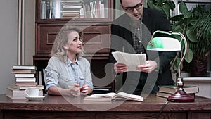 Senior male worker in glasses holding report and explaining tasks to female coworker sitting at the table