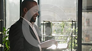 Senior man African American ethnic businessman worker stand in corporate office with laptop in sun sunbeams. Smiling