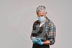 Senior man in apron and protective face mask using digital tablet while standing against gray background