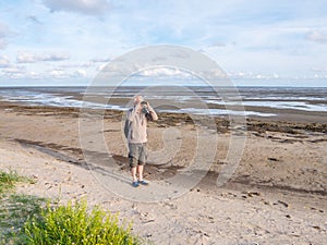 Senior man with binoculars on beach of nature reserve Boschplaat on island Terschelling, Netherlands at low tide of Waddensea