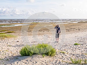 Senior man with binoculars looking at dried out boat on tidal flats at low tide of Waddensea from beach of Boschplaat on