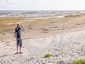 Senior man with binoculars looking at tidal flats at low tide of Waddensea from beach of Boschplaat on Terschelling, Netherlands