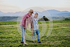 Senior man with broken leg and crutches standing in the the middle of meadow.