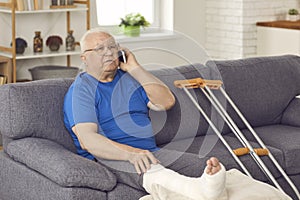 Senior man with broken leg sitting on couch and talking to doctor or family on mobile photo