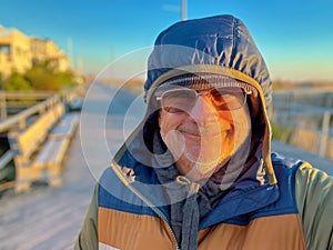 Senior man bundled up in winter coat parker with hood on wearing sunglasses smiling on an empty boardwalk