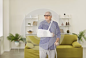 Senior man with a cast and on his broken arm and with a stick standing in the living room at home