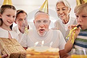 Senior man celebrating his birthday with his family at home, wearing party hats and blowing whistles. Grandpa blowing