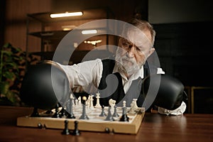 Senior man chess player moving chesspiece on board while wearing in boxer gloves photo