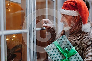 Senior man with a Christmas gift in hand standing in front of the window and knocking on it. Elderly man as Santa Claus