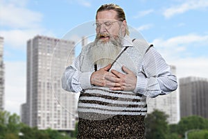 Senior man clutching his chest because of terrible pain standing on city buildings background.