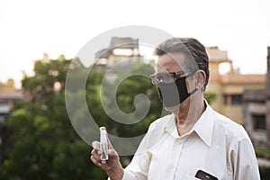Senior man with corona preventive mask showing medicine ampul or vaccine standing on rooftop outdoor. Covid, Lock down and Home