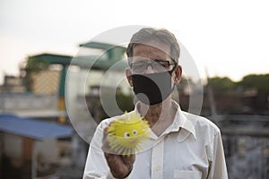 Senior man with corona preventive mask showing a toy model of corona virus standing on rooftop outdoor.
