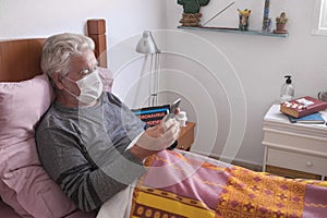 The senior man with COVID-19 lies in bed at home wearing a medical mask. One elderly people at home using cellphone. Quarantine