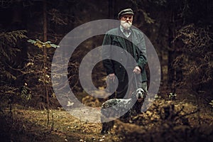 Senior man with dog in a traditional shooting clothing, posing on a dark forest background.