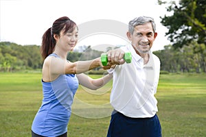 senior man doing exercise with daughter in the nature park,young asian woman train her father using dumbbell