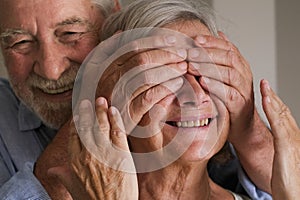 Senior man doing a surprise at his wife mature woman from behind closing her eyes. Happy and joyful elderly people lifestyle home
