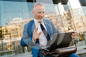 Senior man dressed waving hand and using laptop while sitting by building