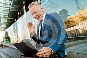 Senior man dressed waving hand and using laptop while sitting by building