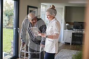 Senior Man In Dressing Gown Using Walking Frame Being Helped By Female Nurse With Digital Tablet