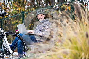 A senior man with electrobike sitting on a bench outdoors in town, using laptop.