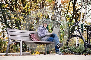 A senior man with electrobike sitting on a bench outdoors in town, using laptop.