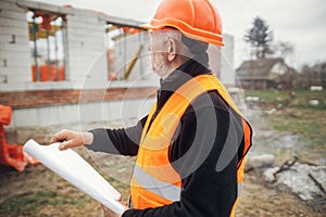 Senior man engineer or construction worker in hardhat looking at blueprints at building new modern house. Male architect or