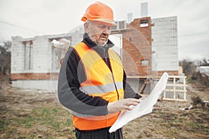 Senior man engineer or construction worker in hardhat looking at blueprints at building new modern house. Male architect or