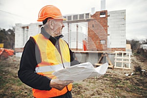 Senior man engineer or construction worker in hardhat looking at blueprints at building new modern house. Male architect or
