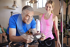 Senior Man Exercising On Cycling Machine Being Encouraged By Female Personal Trainer In Gym