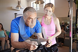 Senior Man Exercising On Cycling Machine Being Encouraged By Female Personal Trainer In Gym