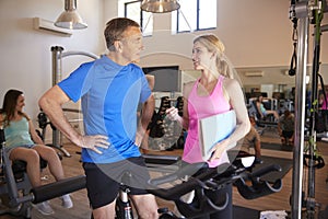 Senior Man Exercising On Cycling Machine Being Encouraged By Female Personal Trainer In Gym