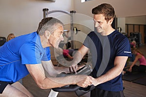 Senior Man Exercising On Cycling Machine Being Encouraged By Personal Trainer In Gym