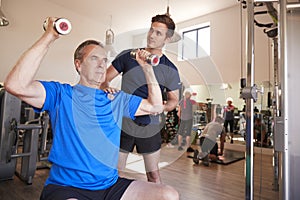 Senior Man Exercising With Weights Being Encouraged By Personal Trainer In Gym
