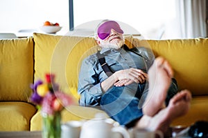 Senior man with eye mask lying on sofa indoors at home, relaxing.