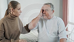 Senior man feel strong headache, touch his head and looks at anxious adult daughter. Worried woman sits on a bed with