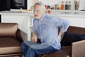 Senior man feeling lower back pain, sitting on armchair