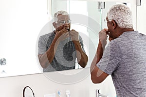 Senior Man Flossing Teeth Looking At Reflection In Bathroom Mirror Wearing Pajamas