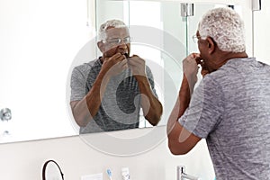 Senior Man Flossing Teeth Looking At Reflection In Bathroom Mirror Wearing Pajamas photo