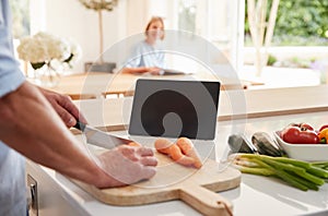 Senior Man Following Recipe On Digital Tablet As Wife Sits At Dining Room Table An Reads Magazine