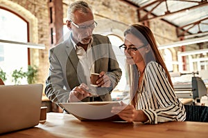 Senior man in formal wear holding coffee cup and explaining something to his young female colleague while working