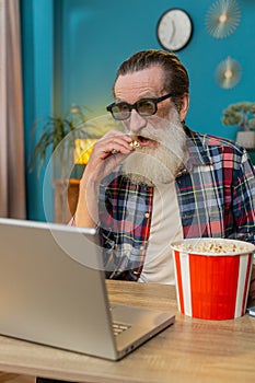 Senior man freelancer taking break from work wearing 3D glasses eating popcorn watching movie film