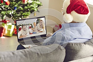 Senior man grandfather in Santa festive hat sitting on sofa and communicating with family online at Christmas