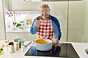 Senior man with grey hair cooking spaghetti at home kitchen pointing thumb up to the side smiling happy with open mouth