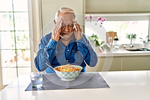 Senior man with grey hair eating pasta spaghetti at home with hand on head for pain in head because stress