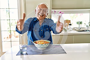 Senior man with grey hair eating pasta spaghetti at home looking at the camera smiling with open arms for hug