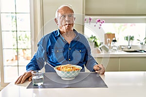Senior man with grey hair eating pasta spaghetti at home making fish face with lips, crazy and comical gesture
