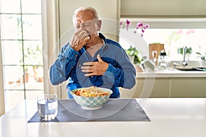 Senior man with grey hair eating pasta spaghetti at home smelling something stinky and disgusting, intolerable smell, holding