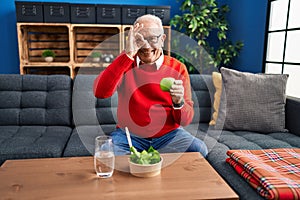 Senior man with grey hair eating salad and green apple smiling happy doing ok sign with hand on eye looking through fingers