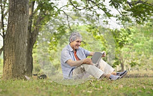 senior man with grey hair holding a book, smiling, sitting near a big tree in forest park