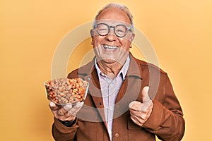 Senior man with grey hair holding fried peanuts smiling happy and positive, thumb up doing excellent and approval sign